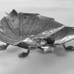 K&ouml;nnte beinhalten: Ein silberfarbener Spielkartenhalter mit f&auml;cherf&ouml;rmigem Design. Der Halter zeigt detaillierte Illustrationen von Spielkarten, die von dekorativen F&uuml;&szlig;en getragen werden. Die Karten sind so angeordnet, dass die Gesichter sichtbar sind.