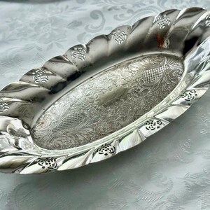 May include: Oval silver serving dish with a decorative, scalloped edge. The dish features an ornate, engraved floral pattern in the center and along the rim. A vintage piece, perfect for serving appetizers or displaying small items.