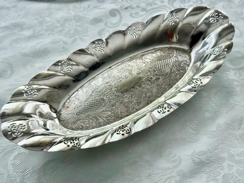 May include: Oval silver serving dish with a decorative, scalloped edge. The dish features an ornate, engraved floral pattern in the center and along the rim. A vintage piece, perfect for serving appetizers or displaying small items.
