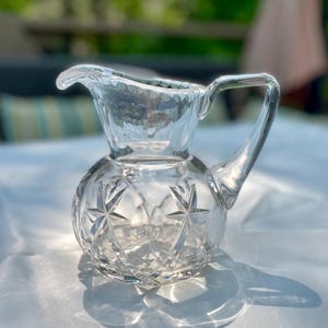 May include: A clear, vintage crystal creamer pitcher with a starburst pattern on the rounded base. The pitcher has a handle and a spout for easy pouring. The glass reflects light, creating a sparkling effect.