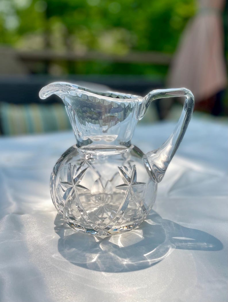 May include: A clear, vintage crystal creamer pitcher with a starburst pattern on the rounded base. The pitcher has a handle and a spout for easy pouring. The glass reflects light, creating a sparkling effect.