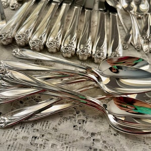May include: A set of vintage silver flatware, including spoons and forks, with ornate floral designs on the handles. The silverware is arranged on a white lace tablecloth.