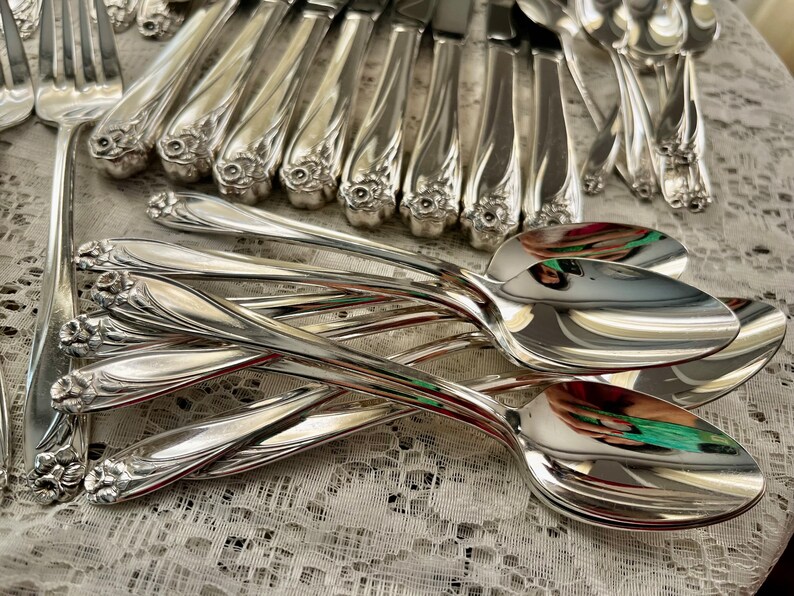 May include: A set of vintage silver flatware, including spoons and forks, with ornate floral designs on the handles. The silverware is arranged on a white lace tablecloth.