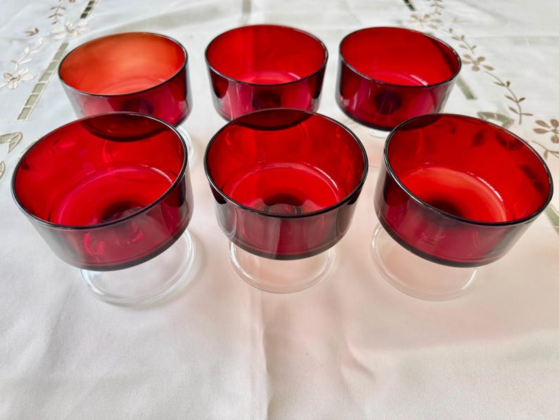 May include: Six clear glass dessert cups with red glass bases. The cups are arranged in two rows of three on a white tablecloth.
