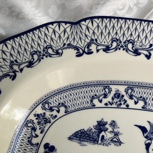 May include: An oval ceramic platter featuring a cream-colored base with intricate dark blue transferware patterns. The design includes a scalloped edge, a lattice border, ornate scrollwork, and a central scene depicting a pagoda and birds.