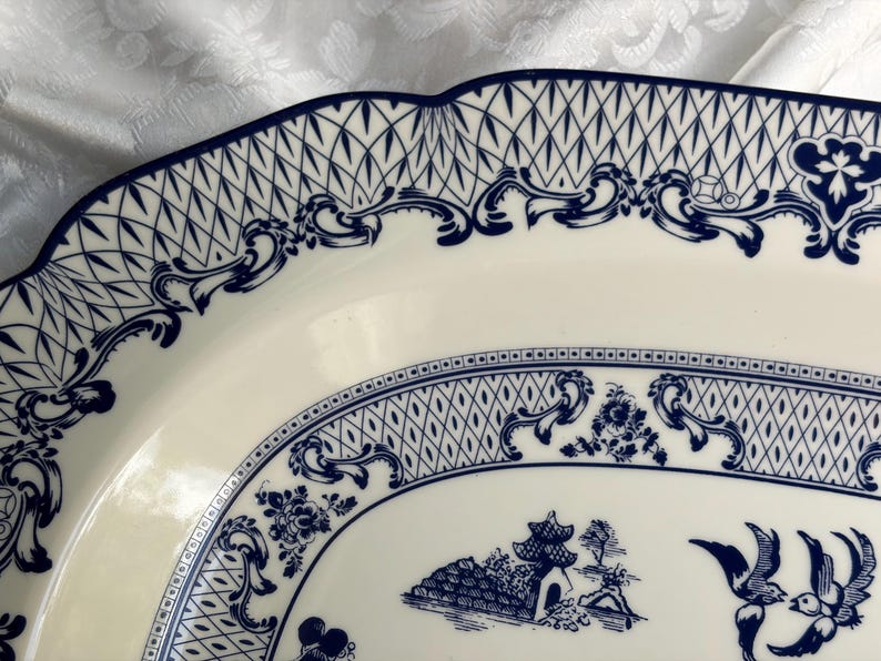 May include: An oval ceramic platter featuring a cream-colored base with intricate dark blue transferware patterns. The design includes a scalloped edge, a lattice border, ornate scrollwork, and a central scene depicting a pagoda and birds.