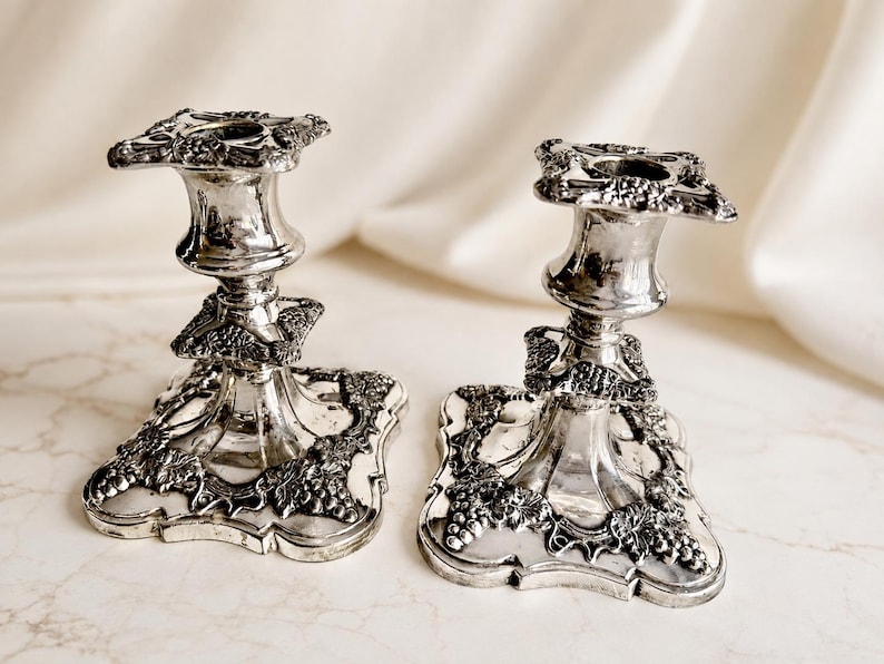 May include: A pair of silver-toned metal candlesticks featuring a square base and top, both decorated with embossed grape and vine leaf motifs. The candlesticks have a polished, reflective finish and a flared stem design with ornate detailing.