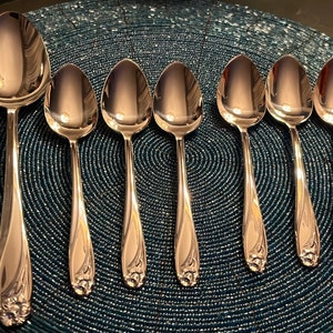 May include: A set of seven polished silver spoons arranged on a textured blue surface. The spoons have a decorative floral design at the base of the handle. The spoons vary in size, with one larger spoon and six smaller spoons.