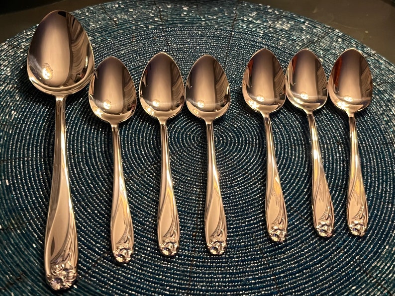 May include: A set of seven polished silver spoons arranged on a textured blue surface. The spoons have a decorative floral design at the base of the handle. The spoons vary in size, with one larger spoon and six smaller spoons.