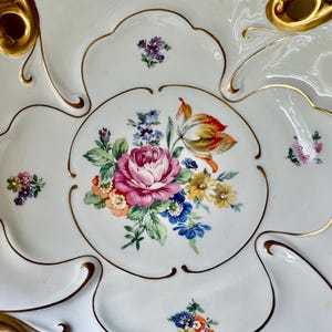 Puede incluir: Una fuente de porcelana blanca con un dise&ntilde;o floral. El centro presenta una gran rosa rosa rodeada de otras flores coloridas. Los bordes est&aacute;n adornados con ribetes y detalles dorados.