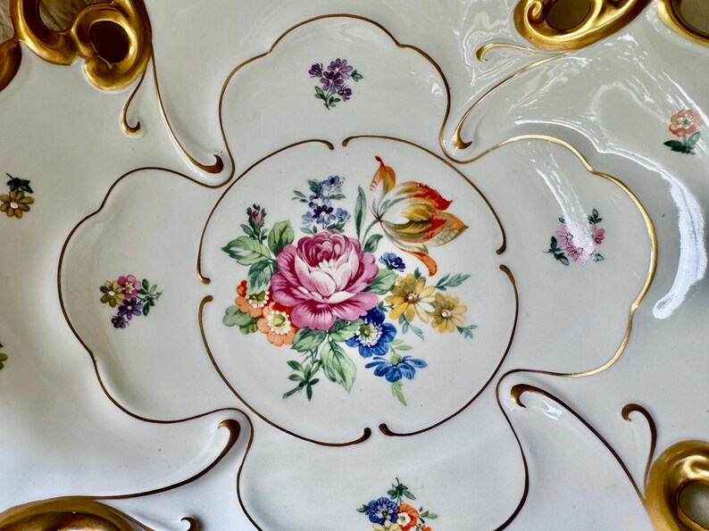 Puede incluir: Una fuente de porcelana blanca con un dise&ntilde;o floral. El centro presenta una gran rosa rosa rodeada de otras flores coloridas. Los bordes est&aacute;n adornados con ribetes y detalles dorados.
