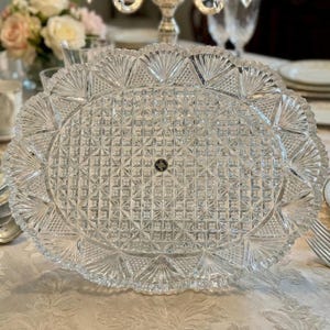 May include: A clear, oval-shaped crystal serving platter with a detailed, geometric pattern. The platter features a scalloped edge with fan-like designs. A small, black and gold emblem is visible in the center. This elegant piece is suitable for serving food at a formal event.