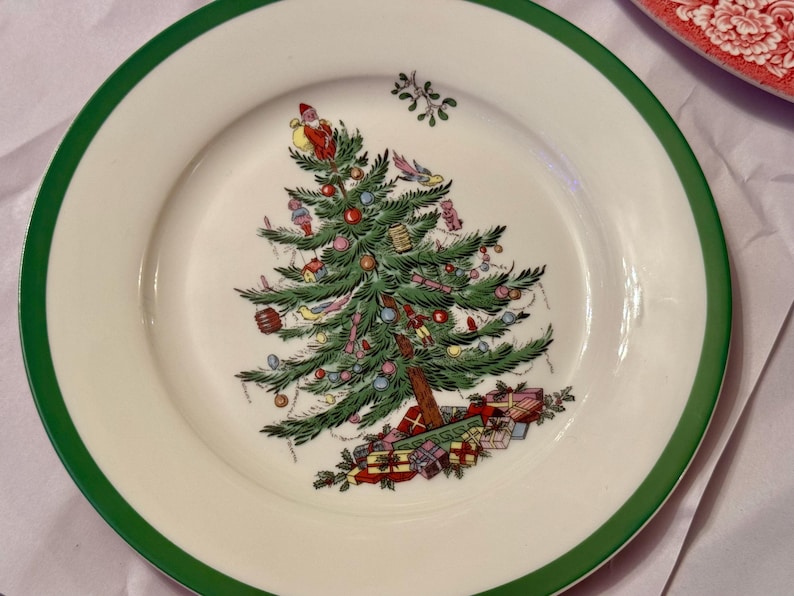 以下が含まれることがあります： 緑色の縁取りのある白いセラミック製のプレートには、赤、緑、金色のオーナメントが飾られたクリスマスツリーのデザインが施されています。ツリーはプレゼントとヤドリギの枝で囲まれています。