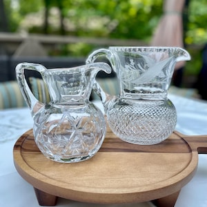 May include: Two clear glass pitchers with intricate cut crystal designs. One pitcher features a diamond pattern, while the other has a floral etched design. They sit on a round wooden serving tray with a handle.