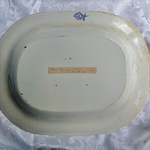 May include: An oval ceramic platter viewed from the underside, featuring a white glazed surface with light brown discoloration along the rim. A blue maker's mark is stamped at the top, and a rectangular paper label with handwritten text is centered.