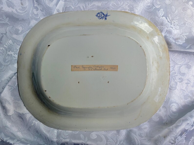 May include: An oval ceramic platter viewed from the underside, featuring a white glazed surface with light brown discoloration along the rim. A blue maker's mark is stamped at the top, and a rectangular paper label with handwritten text is centered.