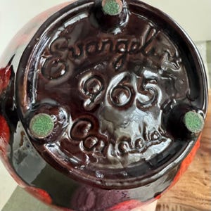 Puede incluir: Primer plano de la base de un jarr&oacute;n de cer&aacute;mica con un esmalte brillante de color marr&oacute;n oscuro. La base presenta el texto en relieve "Evangel" sobre el n&uacute;mero "965" y "Canada". El jarr&oacute;n tiene tres peque&ntilde;as almohadillas de fieltro verde.