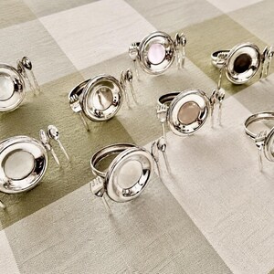 Op de afbeelding: Een set van acht zilverkleurige servetringen, elk voorzien van een cirkelvormig frame met een centrale inzet en decoratieve miniatuur bestekaccenten, waaronder een kleine vork en lepel bevestigd aan de zijkanten van de ring.