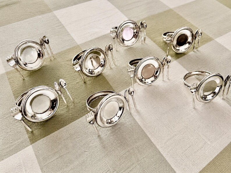 Op de afbeelding: Een set van acht zilverkleurige servetringen, elk voorzien van een cirkelvormig frame met een centrale inzet en decoratieve miniatuur bestekaccenten, waaronder een kleine vork en lepel bevestigd aan de zijkanten van de ring.