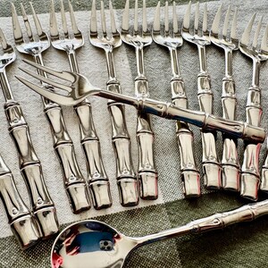 May include: A set of stainless steel flatware featuring a bamboo-inspired handle design. The collection includes multiple forks arranged in a row, one fork placed horizontally across them, and a single spoon positioned at the bottom.