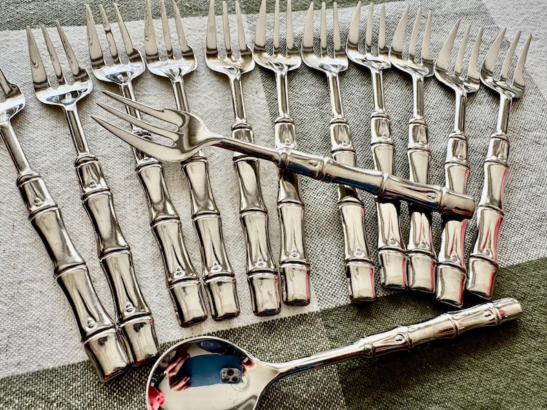 May include: A set of stainless steel flatware featuring a bamboo-inspired handle design. The collection includes multiple forks arranged in a row, one fork placed horizontally across them, and a single spoon positioned at the bottom.