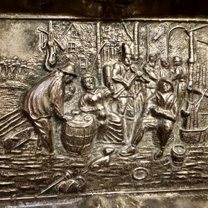 May include: A rectangular silver-toned metal plaque featuring a detailed embossed scene of several figures gathered outdoors. The relief depicts people interacting near a barrel, with trees, a small building, and various tools visible in the background.