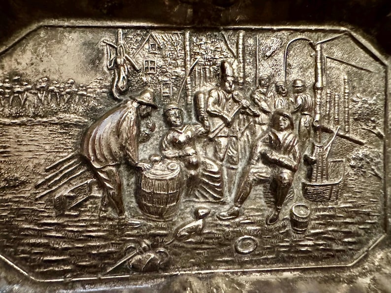 May include: A rectangular silver-toned metal plaque featuring a detailed embossed scene of several figures gathered outdoors. The relief depicts people interacting near a barrel, with trees, a small building, and various tools visible in the background.