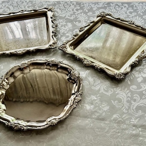 May include: Three silver-coloured serving trays with ornate, scalloped edges. Two are rectangular, and one is oval. The trays are reflective, and the background is a light-coloured fabric with a floral pattern. The trays are empty.