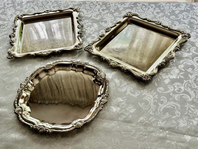 Puede incluir: Tres bandejas de servicio de metal plateado con superficies reflectantes y bordes festoneados ornamentados. Dos de las bandejas son cuadradas con esquinas redondeadas, mientras que la tercera es ovalada. Cada pieza tiene un acabado pulido.