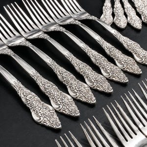 May include: A collection of silver-toned metal forks arranged in rows. Each fork features a detailed floral embossed pattern on the handle, extending from the neck to the base. The tines are long and slender, reflecting light against the dark surface.