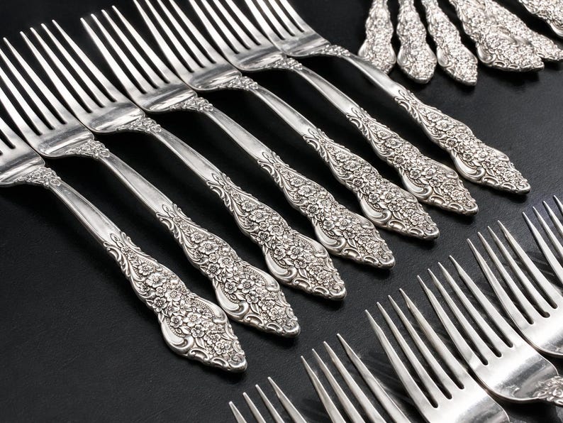 May include: A collection of silver-toned metal forks arranged in rows. Each fork features a detailed floral embossed pattern on the handle, extending from the neck to the base. The tines are long and slender, reflecting light against the dark surface.