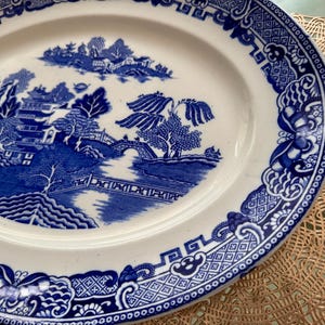 K&ouml;nnte beinhalten: Ein wei&szlig;er ovaler Keramikteller mit einem blauen und wei&szlig;en Blumenmuster. Das Muster zeigt eine Br&uuml;cke, B&auml;ume und eine Pagode. Der Teller hat einen gewellten Rand.