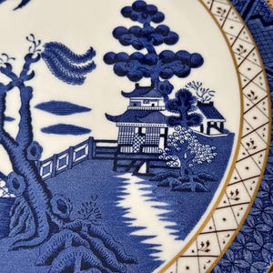 May include: A blue and white porcelain plate with a traditional Chinese design. The plate features a pagoda, trees, and a river. The border of the plate is gold.