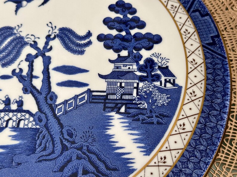 May include: A blue and white porcelain plate with a traditional Chinese design. The plate features a pagoda, trees, and a river. The border of the plate is gold.