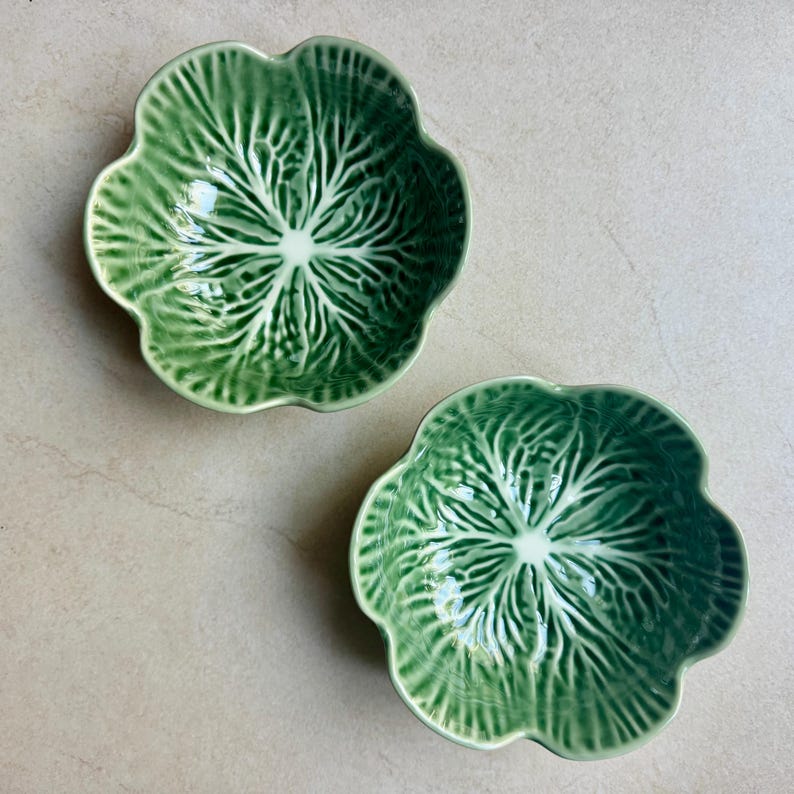 May include: Two ceramic bowls shaped like cabbage leaves, featuring a glossy finish with shades of deep and light green. The bowls have a scalloped edge and a textured surface that mimics the intricate veins and structure of a cabbage leaf.