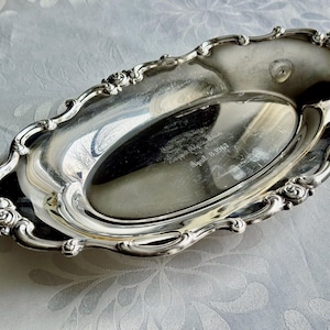 K&ouml;nnte beinhalten: Ein ovales Tablett aus silberfarbenem Metall mit polierter, reflektierender Oberfl&auml;che und einem verzierten, gewellten Rand mit gepr&auml;gten Blumendetails. In der Mitte ist der Text Kappa Alpha Theta und das Datum April 13 1967 eingraviert.