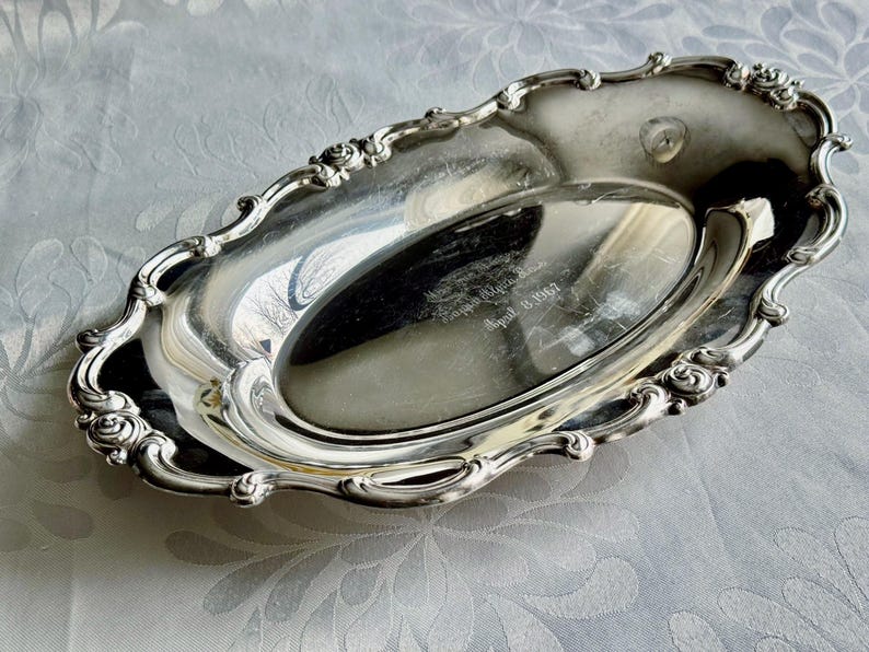K&ouml;nnte beinhalten: Ein ovales Tablett aus silberfarbenem Metall mit polierter, reflektierender Oberfl&auml;che und einem verzierten, gewellten Rand mit gepr&auml;gten Blumendetails. In der Mitte ist der Text Kappa Alpha Theta und das Datum April 13 1967 eingraviert.