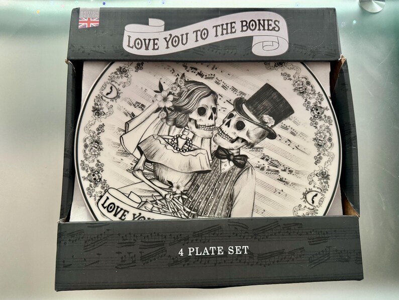 May include: A black and white plate set featuring a skeleton couple in wedding attire. The plate design includes musical notes and the phrase "Love You To The Bones." The box reads "4 PLATE SET" and has a Union Jack flag.