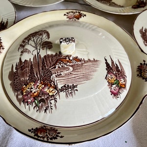 May include: A vintage white ceramic serving dish with a brown and cream floral design. The dish has a lid with a raised knob and a scene of a cottage in a countryside setting.