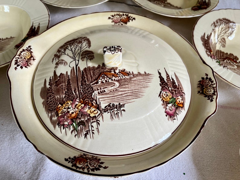 May include: A vintage white ceramic serving dish with a brown and cream floral design. The dish has a lid with a raised knob and a scene of a cottage in a countryside setting.