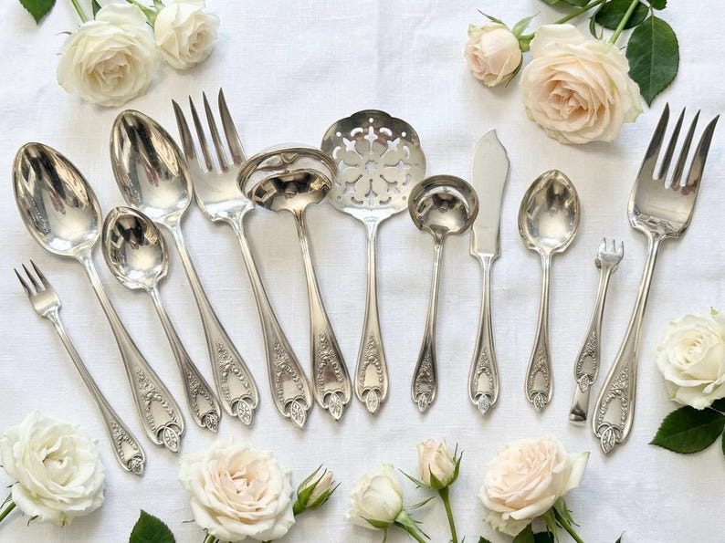 May include: A collection of silver-toned flatware including various spoons, forks, a ladle, a slotted serving spoon, and a knife. Each piece features an ornate, embossed handle design. The set is arranged on a white fabric surface with pale roses.
