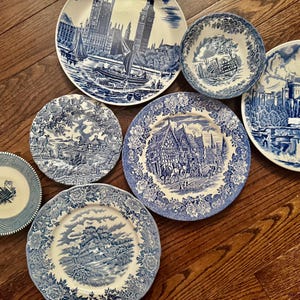 May include: A collection of six ceramic plates and one bowl featuring blue and white transferware patterns. The designs include detailed illustrations of historic buildings, landscapes, and architectural scenes, all rendered in a classic blue ink.