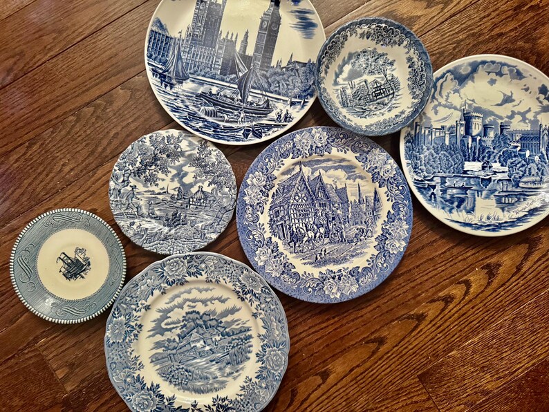 May include: A collection of six ceramic plates and one bowl featuring blue and white transferware patterns. The designs include detailed illustrations of historic buildings, landscapes, and architectural scenes, all rendered in a classic blue ink.