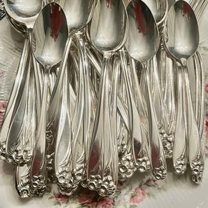 May include: A collection of silver spoons with ornate floral detailing on the handles. The spoons are arranged in a close-up shot, showcasing their reflective surfaces and intricate design. The spoons are likely part of a vintage silverware set.