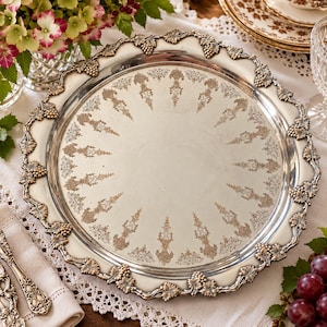 May include: A round silver serving tray featuring an ornate border with raised grape and leaf motifs. The center of the tray is polished silver with a circular pattern of etched, light-colored decorative filigree designs.