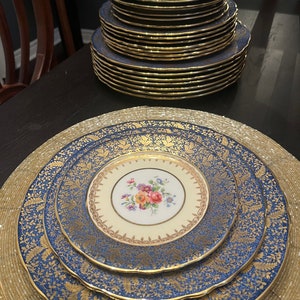 May include: A collection of vintage porcelain plates with a blue and gold floral design. The plates have a cream-colored center with a floral bouquet. The edges are scalloped and trimmed with gold. Several plates are stacked on a dark surface.