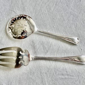 May include: Two silver serving utensils: a slotted spoon and a three-pronged fork. The spoon has a decorative, pierced bowl. Both utensils have heart-shaped details at the end of their handles. The items are on a light gray linen surface.