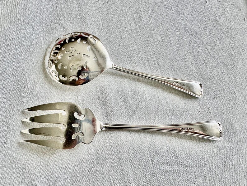 May include: Two silver serving utensils: a slotted spoon and a three-pronged fork. The spoon has a decorative, pierced bowl. Both utensils have heart-shaped details at the end of their handles. The items are on a light gray linen surface.