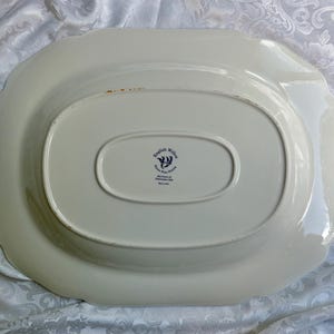 May include: An oval-shaped cream-colored ceramic serving platter viewed from the underside. The center features a dark blue stamp with the text English Willow, Iron Pot Stone, Johnson Bros, Made in England. The edges have a scalloped design.