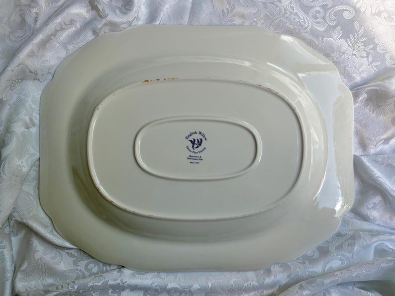 May include: An oval-shaped cream-colored ceramic serving platter viewed from the underside. The center features a dark blue stamp with the text English Willow, Iron Pot Stone, Johnson Bros, Made in England. The edges have a scalloped design.